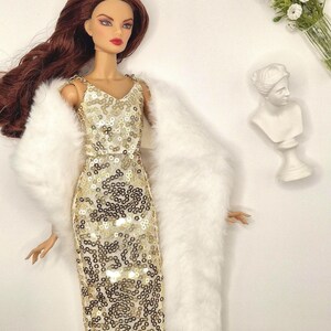 May include: A fashion doll wearing a gold sequined dress and a white faux fur wrap. The doll has long auburn hair and is posed with a white bust sculpture and a small bouquet of white flowers in the background.