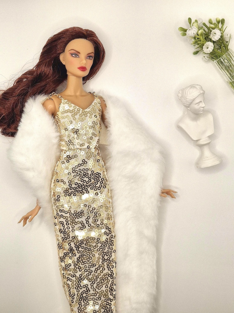 May include: A fashion doll wearing a gold sequined dress and a white faux fur wrap. The doll has long auburn hair and is posed with a white bust sculpture and a small bouquet of white flowers in the background.
