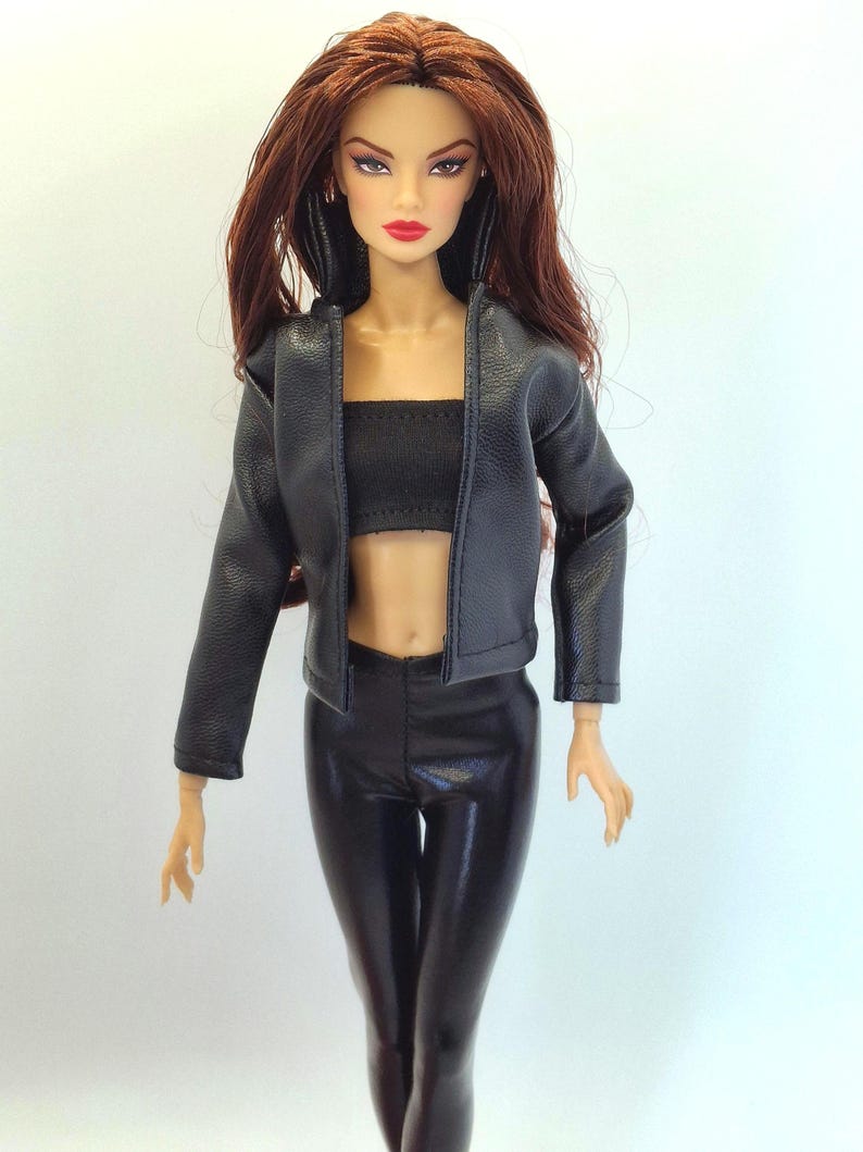 May include: A fashion doll with long auburn hair, wearing a black leather jacket, a black tube top, and black shiny pants. The doll has red lipstick and is posed against a white background.