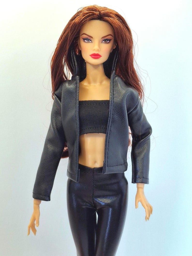 May include: A fashion doll with long auburn hair, wearing a black leather jacket, a black tube top, and black shiny pants. The doll has red lipstick and is posed against a plain background. The doll's outfit is a modern style.