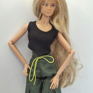May include: A fashion doll with long blonde hair, wearing a black sleeveless crop top and olive green cargo pants featuring a bright neon yellow drawstring waist tie. The doll has painted facial features, including winged eyeliner and freckles.