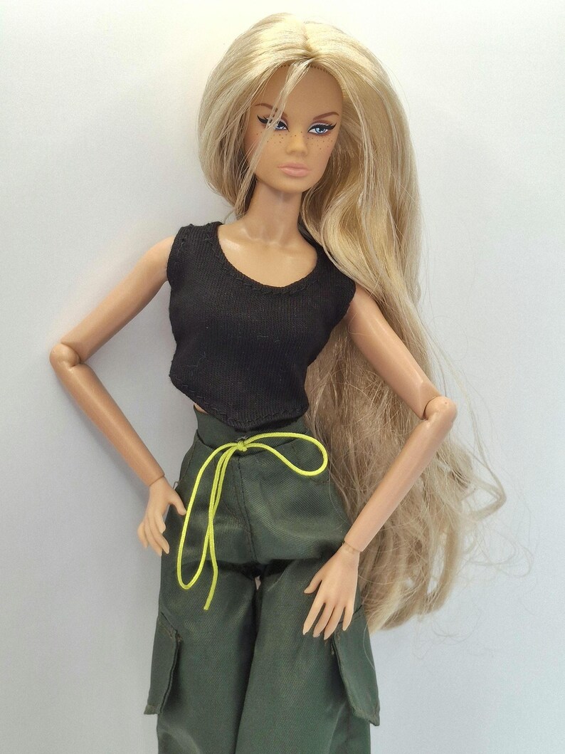 May include: A fashion doll with long blonde hair, wearing a black sleeveless crop top and olive green cargo pants featuring a bright neon yellow drawstring waist tie. The doll has painted facial features, including winged eyeliner and freckles.