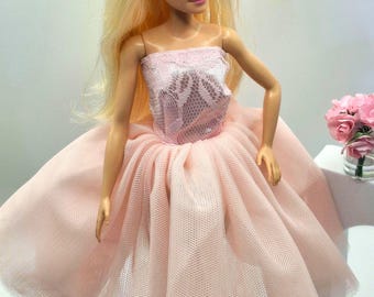 May include: A blonde doll with blue eyes wearing a strapless dress featuring a pale pink lace bodice and a voluminous, layered tulle skirt. The doll has small white flower-shaped earrings and is positioned near a small glass vase of pink roses.