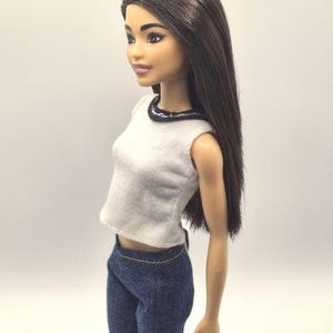 May include: A fashion doll with long brown hair, wearing a white sleeveless top with a black trim and blue denim pants. The doll has a neutral expression and is posed in a side profile.