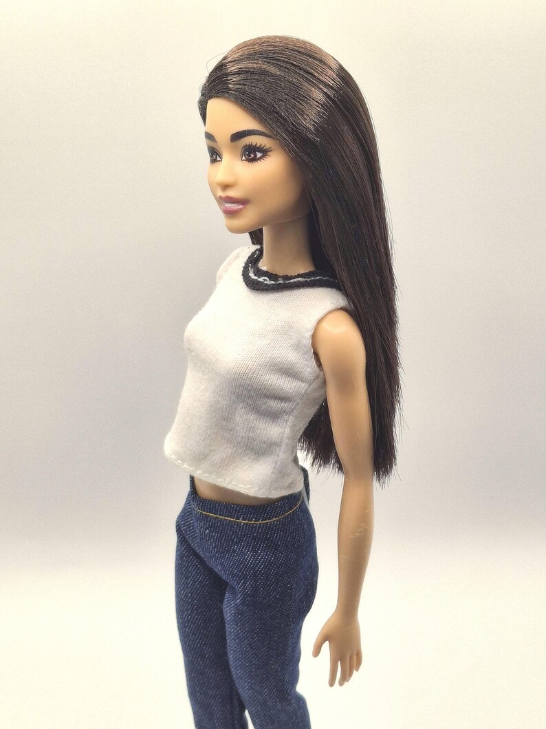 May include: A fashion doll with long brown hair, wearing a white sleeveless top with a black trim and blue denim pants. The doll has a neutral expression and is posed in a side profile.