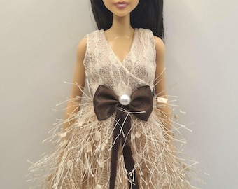 May include: A doll with long dark hair and pearl stud earrings wearing a sleeveless beige dress. The dress features a lace bodice and a skirt made of textured fringe fabric, accented at the waist with a brown ribbon bow and a central pearl.