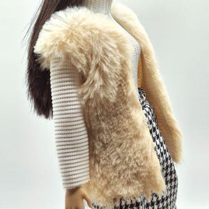 May include: A doll wearing a cream-colored faux fur vest over a white ribbed sweater. The doll is also wearing a black and white houndstooth skirt. The doll has long brown hair.