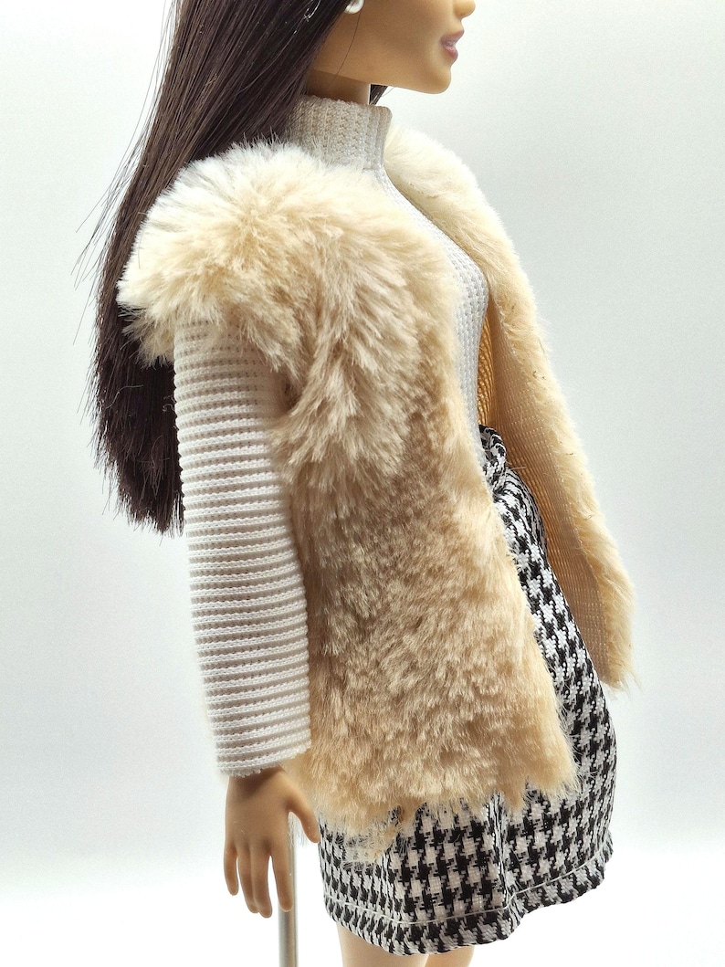 May include: A doll wearing a cream-colored faux fur vest over a white ribbed sweater. The doll is also wearing a black and white houndstooth skirt. The doll has long brown hair.