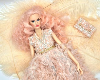 May include: A fashion doll with pink hair and blue eyes, wearing a pink and white dress with a gold belt. A small matching purse is next to the doll. The doll is lying on a bed of feathers.