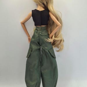May include: A doll with long blonde hair wearing a black sleeveless crop top and olive green cargo pants with side pockets and elastic cuffs at the ankles. The doll is viewed from the back, showing the fit of the clothing and hair length.