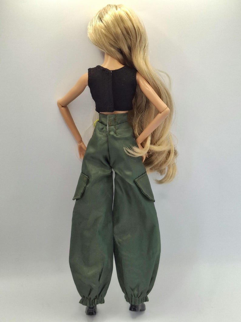 May include: A doll with long blonde hair wearing a black sleeveless crop top and olive green cargo pants with side pockets and elastic cuffs at the ankles. The doll is viewed from the back, showing the fit of the clothing and hair length.