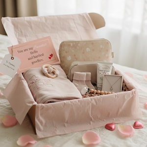 May include: A pink gift box filled with baby items, including a blanket, socks, a teething ring, and a necklace. A card reads "You are my sweetest Hello and hardest goodbye." The box is lined with pink tissue paper and surrounded by rose petals.