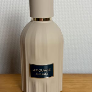 Puede incluir: Un frasco de perfume beige con una tapa a juego. La botella tiene un diseño texturizado y acanalado y una banda dorada cerca de la parte superior. Una etiqueta azul oscuro con letras doradas dice "AMOUAGE OUTLANDS". La botella está sobre una superficie de madera.