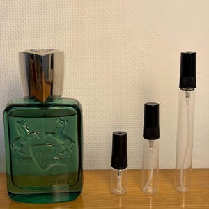 May include: A green glass perfume bottle with a silver cap and a shield design with two horses. There are also three clear glass vials with black caps, used for perfume samples.