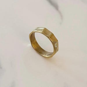 May include: A gold ring with a faceted design. The ring has a geometric shape with multiple flat surfaces, creating a textured appearance. The ring is set against a white marble background.