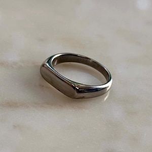 May include: A silver ring with a rectangular face and rounded edges. The ring has a smooth, polished finish and a simple, modern design. The jewellery is likely made of stainless steel or a similar metal.