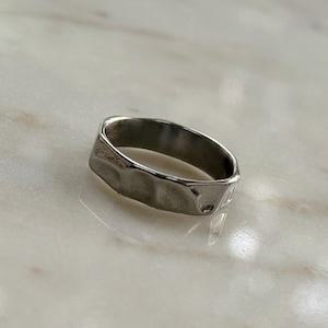 May include: A silver ring with a textured, hammered finish. The ring has an irregular shape, with a band approximately 0.6 cm wide. The surface reflects light, highlighting the uneven texture and creating a subtle, metallic sheen.