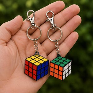 Peut inclure: Deux porte-clés en forme de cubes puzzles miniatures avec des motifs de grille colorés. Un cube présente des faces jaune, bleue et rouge, tandis que l'autre montre des faces verte, orange et blanche. Chaque porte-clés inclut un anneau en métal argenté.