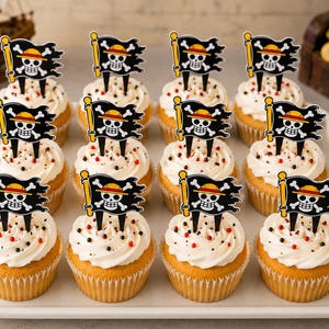 May include: A set of twelve cupcakes arranged on a white rectangular tray, each topped with a black pirate flag topper featuring a white skull and crossbones wearing a yellow straw hat with a red band, and white frosting with small sprinkles.