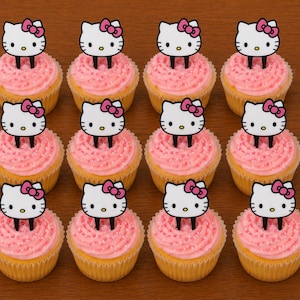 Puede incluir: Doce cupcakes con glaseado rosa y adornos de Hello Kitty. Los cupcakes están sobre una superficie marrón. Cada cupcake tiene una base amarilla, glaseado rosa y una cara de Hello Kitty con un lazo rosa.