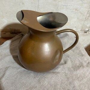 Puede incluir: Una jarra vintage de color cobre con un cuerpo redondeado y un asa curvada. La jarra tiene un pico con un dise&ntilde;o &uacute;nico y asim&eacute;trico. El metal tiene un aspecto ligeramente envejecido, lo que sugiere que es un art&iacute;culo antiguo o vintage.
