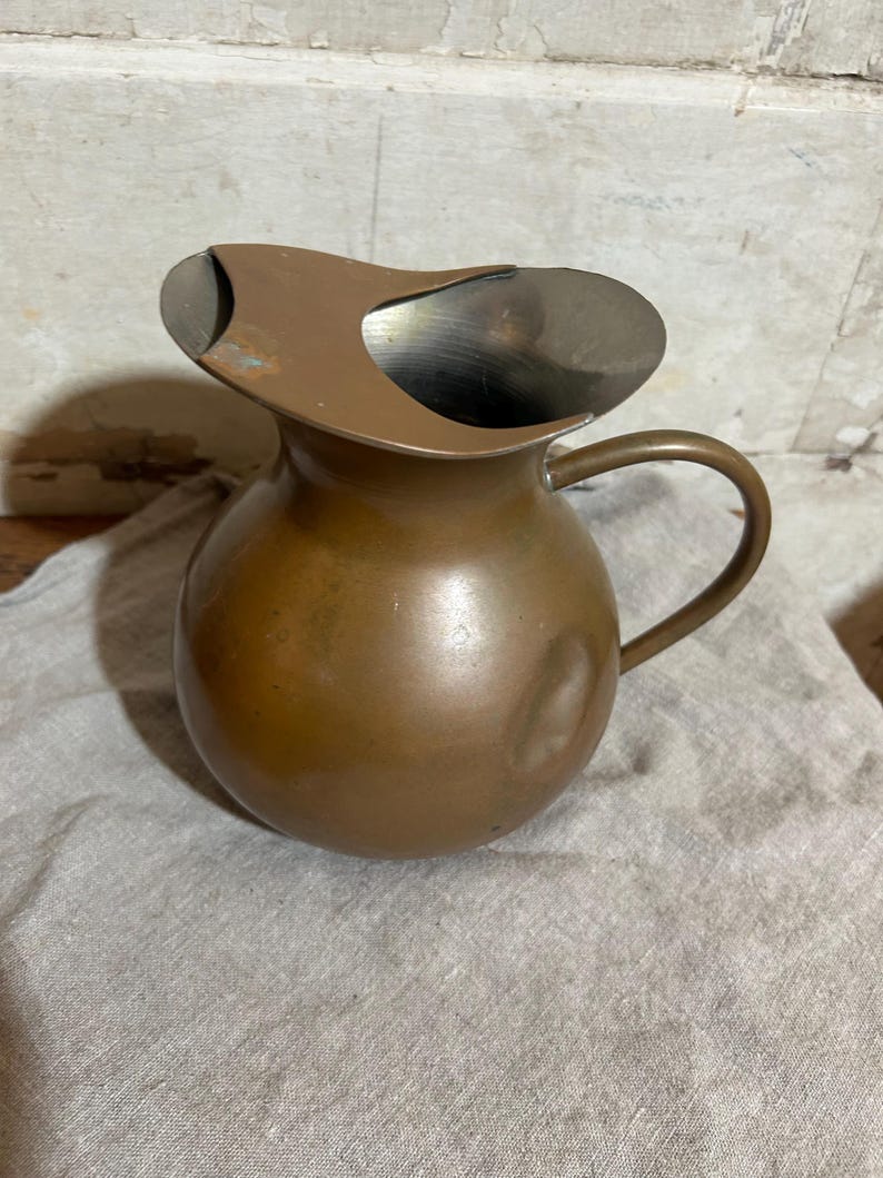 Puede incluir: Una jarra vintage de color cobre con un cuerpo redondeado y un asa curvada. La jarra tiene un pico con un dise&ntilde;o &uacute;nico y asim&eacute;trico. El metal tiene un aspecto ligeramente envejecido, lo que sugiere que es un art&iacute;culo antiguo o vintage.