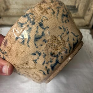May include: A square ceramic bowl with a beige background and blue floral patterns. The bowl has a slightly cracked, aged appearance. The bowl is held by a person's hand.