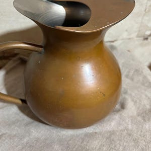 Puede incluir: Una jarra de cobre vintage con un cuerpo redondeado y un pico curvo. La jarra tiene un asa en el lateral y un interior met&aacute;lico brillante. El cobre tiene un color marr&oacute;n c&aacute;lido con algunos signos de edad y uso.