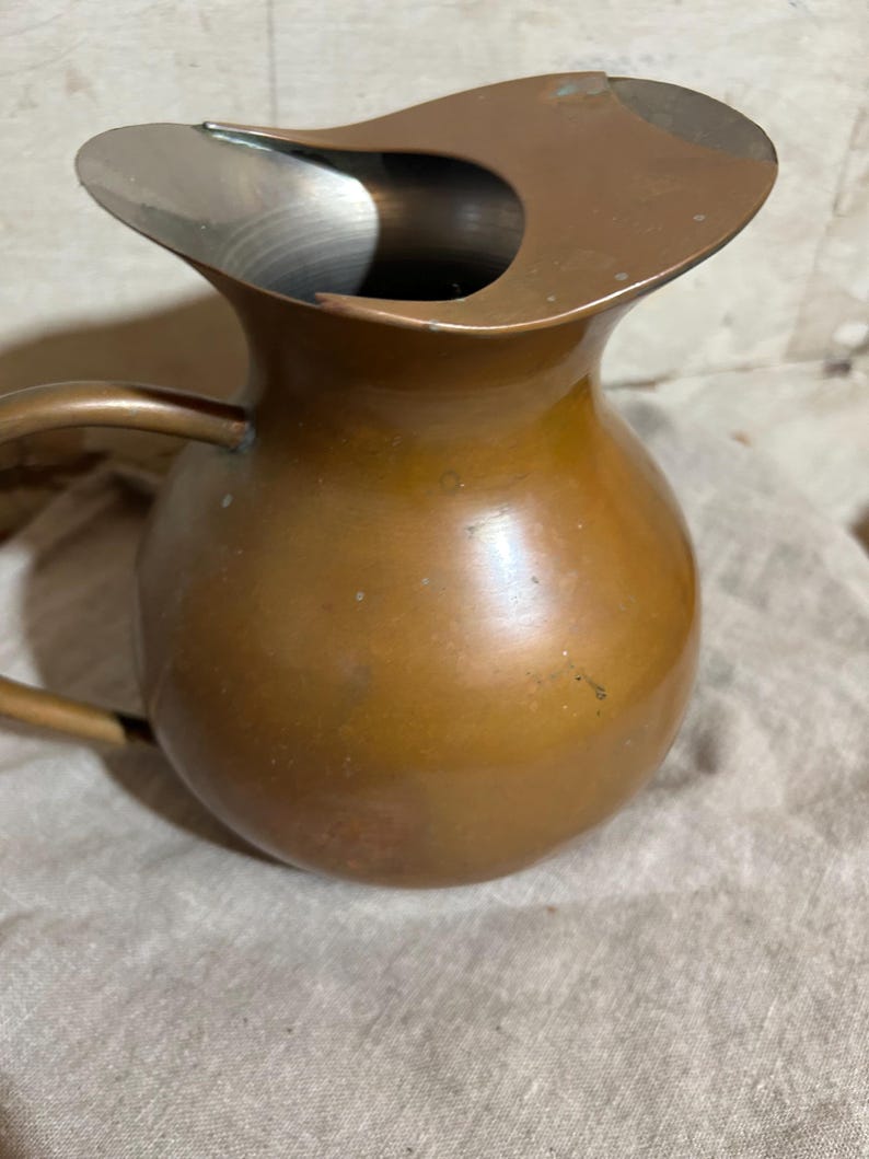 Puede incluir: Una jarra de cobre vintage con un cuerpo redondeado y un pico curvo. La jarra tiene un asa en el lateral y un interior met&aacute;lico brillante. El cobre tiene un color marr&oacute;n c&aacute;lido con algunos signos de edad y uso.