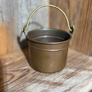 May include: A small, cylindrical copper bucket featuring a smooth, weathered finish and a thin, arched brass handle attached to the sides with metal rivets. The interior of the bucket is dark, and the exterior shows a warm, metallic brown patina.