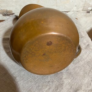 Puede incluir: Una jarra de cobre vintage con un cuerpo redondeado y un asa peque&ntilde;a. La jarra tiene una p&aacute;tina c&aacute;lida y envejecida, con desgaste y decoloraci&oacute;n visibles, lo que sugiere su edad. La jarra est&aacute; sobre un pa&ntilde;o de lino gris claro.