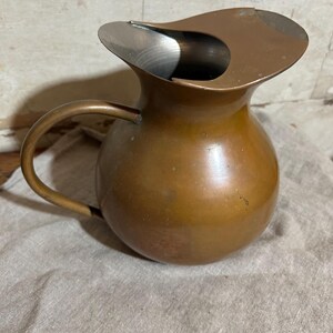 Puede incluir: Una jarra de color cobre con un asa curvada y un dise&ntilde;o de boquilla &uacute;nico. La jarra tiene un cuerpo redondeado y un interior met&aacute;lico brillante. Se encuentra sobre una tela de color neutro.