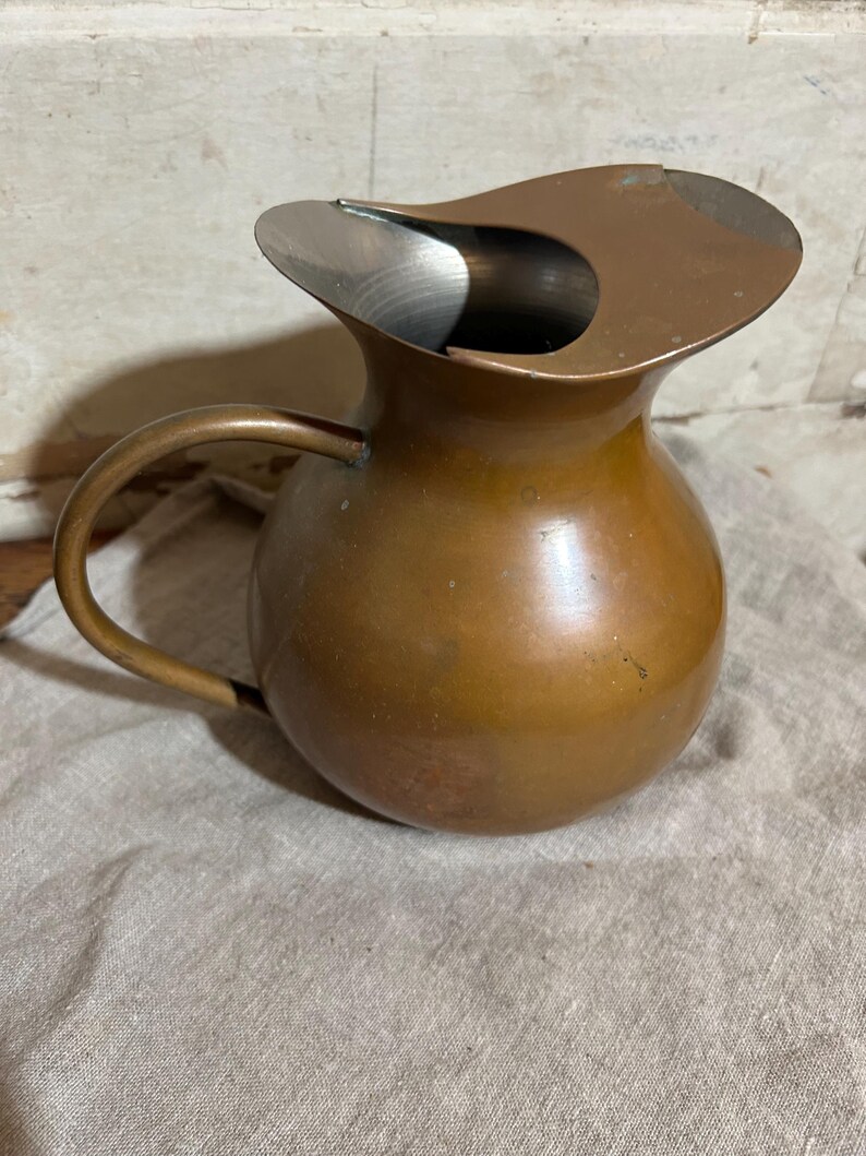 Puede incluir: Una jarra de color cobre con un asa curvada y un dise&ntilde;o de boquilla &uacute;nico. La jarra tiene un cuerpo redondeado y un interior met&aacute;lico brillante. Se encuentra sobre una tela de color neutro.