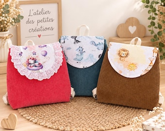 Op de afbeelding: Drie rugzakken van corduroy in rood, donkerblauw en bruin. Elke tas heeft een klep met een unieke print: een eenhoorn met ballonnen, babydinosaurussen en een leeuw met een zon. De rode tas is voorzien van een witte kanten rand langs de klep.