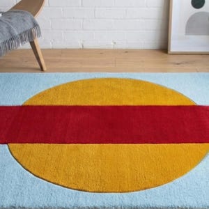 May include: A rectangular rug with a light blue background, featuring a large yellow circle intersected by a red rectangle. The rug is placed on a wooden floor, with a chair and plants in the background.