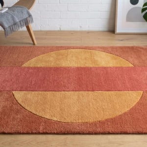 May include: A rectangular rug with a geometric design. The rug features a large, orange-yellow circle bisected by a rectangular band of reddish-orange. The rug is made of a soft, textured material and is placed on a wooden floor.