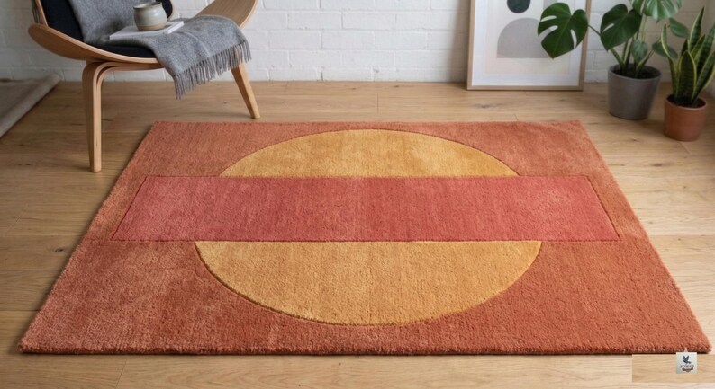 May include: A rectangular rug with a geometric design. The rug features a large, orange-yellow circle bisected by a rectangular band of reddish-orange. The rug is made of a soft, textured material and is placed on a wooden floor.