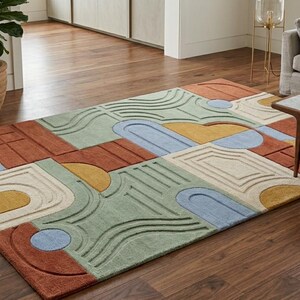 Pode incluir: Um tapete retangular com um padrão geométrico multicolorido e texturas em relevo. O design inclui formas em terracota, verde sálvia, amarelo mostarda, creme e azul claro, como arcos, círculos e linhas, dispostas numa grelha.