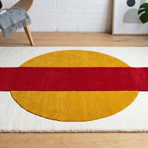 May include: A rectangular rug with a white background, featuring a large yellow circle intersected by a red rectangle. The rug is placed on a wooden floor, with a chair and plants in the background.