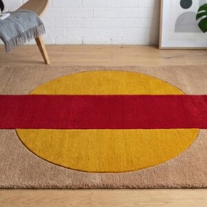 May include: A rectangular area rug with a geometric design. The rug features a large yellow circle with a red rectangular stripe across the center. The rug has a tan border and is placed on a wooden floor.