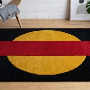 May include: A rectangular black rug with a large yellow circle and a red rectangle across the center. The rug is on a wooden floor, with a chair and plants in the background.