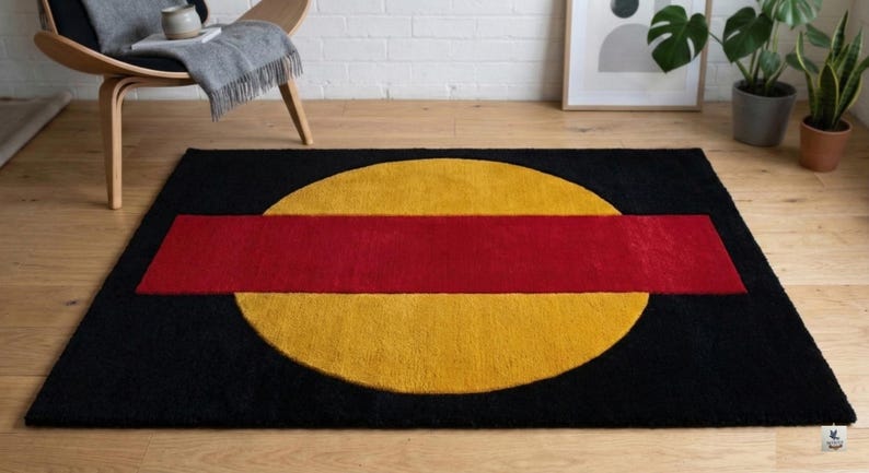 May include: A rectangular black rug with a large yellow circle and a red rectangle across the center. The rug is on a wooden floor, with a chair and plants in the background.