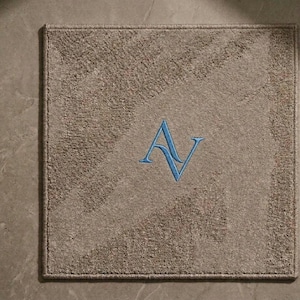 May include: Square, beige rug with a textured pattern and a blue embroidered "AV" monogram. The rug has a darker border. It is placed on a light-coloured surface. The rug is likely used as a decorative floor covering.
