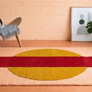 May include: A rectangular rug with a peach-colored background, featuring a large yellow circle and a red rectangular stripe across the center. The rug is placed on a wooden floor, with a chair and artwork in the background.