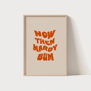 May include: A beige framed print with the text "NOW THEN MARDY BUM" in orange.