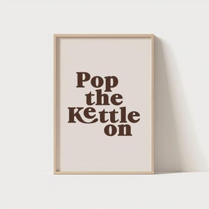 以下が含まれることがあります： 「Pop the Kettle On」という茶色の文字が書かれたフレーム付きプリント。テキストはクリーム色の背景に配置され、明るい茶色の木製フレームに収められています。キッチンやダイニングエリア向けの装飾的なアートワークです。