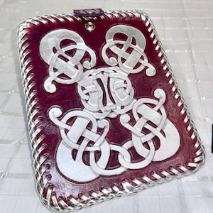Pode incluir: Uma capa de couro bordô com um design de nó celta prateado. A capa tem uma borda prateada costurada e um fecho de pressão roxo. O design é gravado no couro, criando um efeito texturizado.