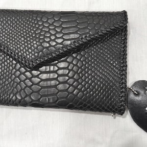 以下が含まれることがあります： 黒いクラッチバッグ。ヘビ革模様と編み込みエッジが特徴です。フラップ開閉とジッパーが付いています。白い文字が書かれた黒いハート型のタグが付属しています。