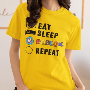 Puede incluir: Camiseta amarilla de manga corta con el texto "EAT SLEEP ROBLOX REPEAT" en negro, azul y rojo. La camiseta presenta una cama, un logotipo de Roblox y un gr&aacute;fico de flecha circular. La camiseta es de material suave.