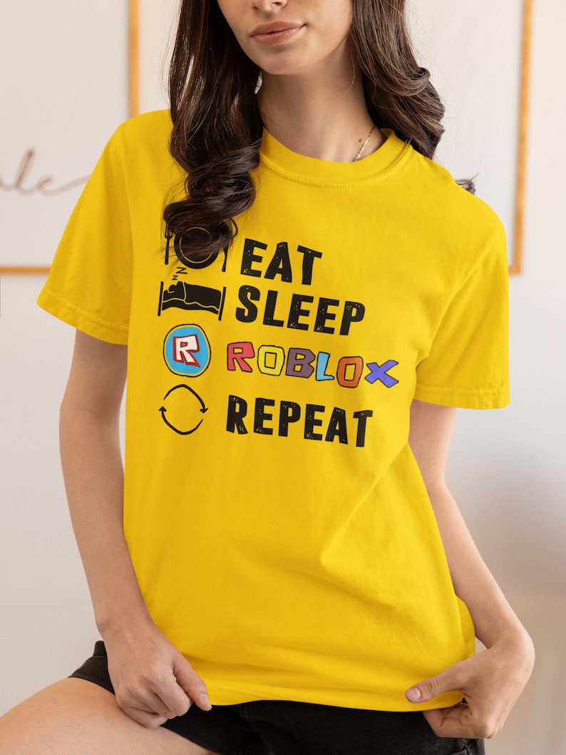 Puede incluir: Camiseta amarilla de manga corta con el texto "EAT SLEEP ROBLOX REPEAT" en negro, azul y rojo. La camiseta presenta una cama, un logotipo de Roblox y un gr&aacute;fico de flecha circular. La camiseta es de material suave.
